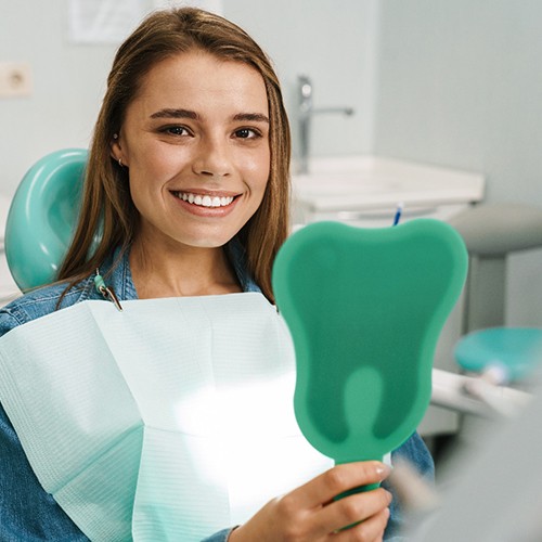 Young woman smiling while holding tooth-shaped mirror in dental chair
